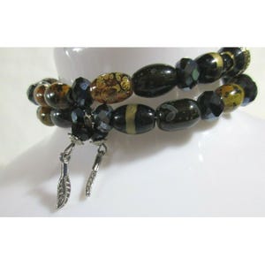 May include: Two beaded bracelets with a variety of dark-colored beads, including black, brown, and gold. The bracelets are adorned with silver-colored charms. The beads have unique patterns and textures, creating a stylish accessory.