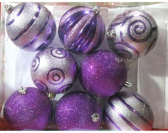 May include: A set of eight Christmas ornaments in a clear plastic container. The ornaments are a mix of silver and purple, with glitter and swirl designs. Some have a shiny finish, while others are covered in glitter.