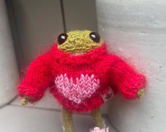 May include: A small green crocheted frog wearing a red knitted sweater with a pink heart design and pink shoes.