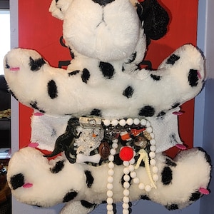 May include: A plush Dalmatian dog with black spots and a black nose, mounted on a red background. The dog's belly is adorned with various small trinkets, including beads, a football charm, and a red pom-pom.