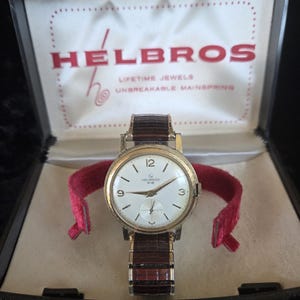 Op de afbeelding: Vintage Helbros horloge in de originele doos. Het horloge heeft een goudkleurige kast, een witte wijzerplaat met zwarte cijfers en een bruine leren band. De doos is zwart met een crèmekleurig interieur en het Helbros logo.