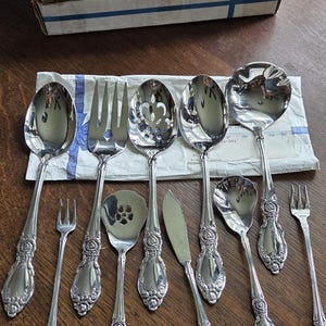 May include: A set of ten silver-toned stainless steel utensils featuring ornate floral embossed handles. The collection includes various spoons, forks, a butter knife, and a ladle, arranged on a white paper wrapper with a blue stripe design.