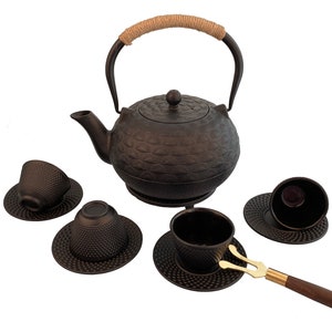 May include: A black cast iron teapot with a brown rope handle and four matching cups and saucers. The teapot and cups have a textured, embossed design. A gold metal tea tong is included.