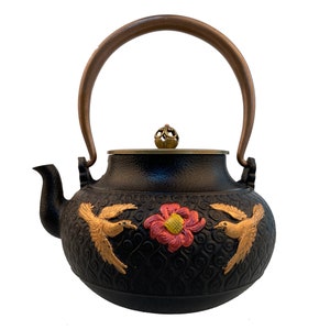 May include: Black cast iron teapot with a brown handle and gold accents. The teapot features a floral design with two birds on either side.