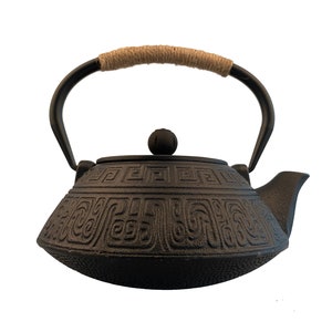 May include: Black cast iron teapot with a rope handle and intricate geometric designs. The teapot has a round lid with a knob on top.
