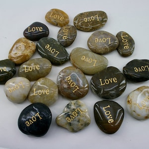 May include: A collection of smooth, natural stones with the word "Love" inscribed in gold lettering. The stones are various shades of brown, gray, and white.