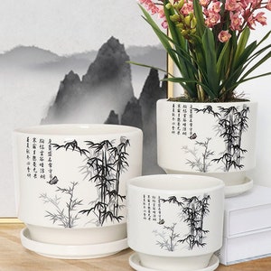 May include: Three white ceramic planters with black bamboo and butterfly designs. The planters have a matching saucer and feature Chinese calligraphy. The largest planter is holding a pink orchid plant.