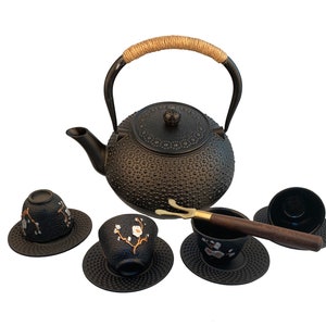 May include: Black cast iron teapot with a brown rope handle and four matching cups and saucers with a floral design. The set also includes a bamboo tea scoop.