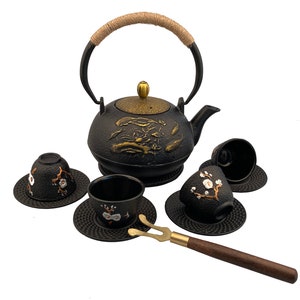 May include: Black cast iron teapot with a gold floral design and a matching set of four teacups and saucers. The teapot has a rope handle and a small strainer. The teacups and saucers have a floral design and a textured surface. A wooden tea tong is included.