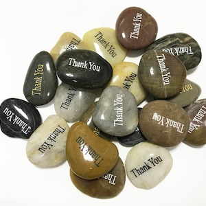 May include: A collection of smooth, gray, brown, and white stones with the words "Thank You" written on them in black and gold lettering.