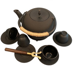 May include: Black cast iron teapot with a brown rope handle and four matching cups and saucers. The teapot and cups have a textured, dimpled surface.