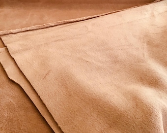 Puede incluir: Un trozo de piel de ante de color marrón claro con una textura suave, mate y ligeramente aterciopelada. El material está doblado, mostrando los bordes crudos y sin rematar, con un tono beige cálido y uniforme en toda la superficie.