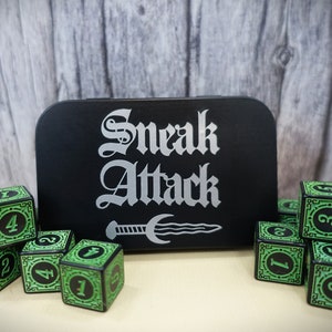 May include: A black metal box with the words "Sneak Attack" in silver lettering and a silver dagger graphic. The box is surrounded by green and black dice with numbers 1, 2, 3, and 4.