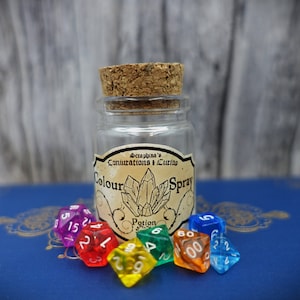 Könnte beinhalten: Eine kleine Glasflasche mit einem Korkverschluss und einem Etikett mit der Aufschrift "Seraphina's Conjuratons + Curios Colour Spray Potion". Die Flasche ist von sechs bunten Würfeln umgeben.