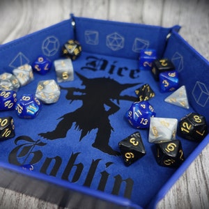 Op de afbeelding: Een blauwe vilten dobbelsteenbak met een zwart silhouet van een goblin en de woorden "Dice Goblin" in gouden letters. De bak is gevuld met verschillende gekleurde dobbelstenen, waaronder zwart, wit en blauw.