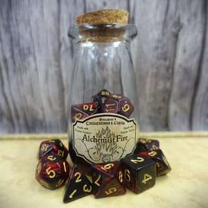 Könnte beinhalten: Ein Glasgefäß mit einem Korkverschluss, gefüllt mit roten und schwarzen Würfeln. Das Gefäß trägt ein Etikett mit der Aufschrift "Seraphina's Conjuratons & Curios Alchemist Fire Potion".