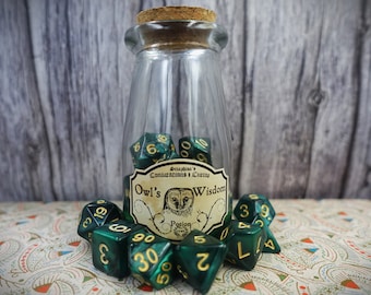 Może przedstawiać: Szklany słoik z korkową pokrywką wypełniony zielonymi i złotymi kostkami do gry. Słoik ma etykietę z napisem "Owl's Wisdom Potion".