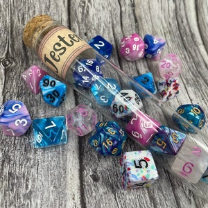 May include: A glass test tube filled with colorful dice, with the label "Tester" on the side. The dice are a mix of blue, pink, and white with black and gold numbers. There are also several dice scattered around the test tube.