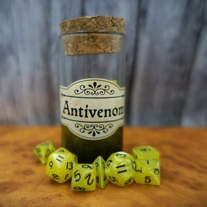May include: A glass bottle with a cork stopper and a label that reads "Antivenom". The bottle is filled with a green liquid. There are six yellow dice with black numbers on them in front of the bottle.