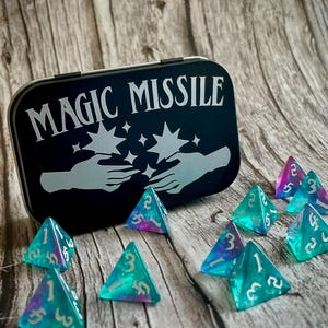 K&ouml;nnte beinhalten: Eine schwarze Metalldose mit den Worten "MAGIC MISSILE" in silbernen Lettern. Die Dose ist ge&ouml;ffnet und enth&auml;lt 10 schimmernde blaue und violette dreieckige W&uuml;rfel mit wei&szlig;en Zahlen.