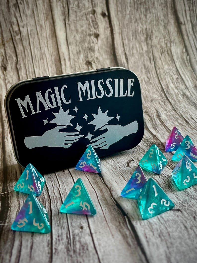 K&ouml;nnte beinhalten: Eine schwarze Metalldose mit den Worten "MAGIC MISSILE" in silbernen Lettern. Die Dose ist ge&ouml;ffnet und enth&auml;lt 10 schimmernde blaue und violette dreieckige W&uuml;rfel mit wei&szlig;en Zahlen.