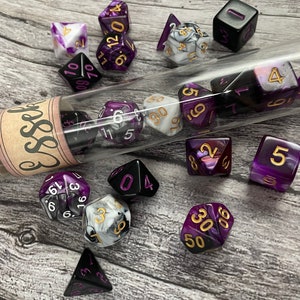 May include: A glass test tube filled with purple and black dice with gold numbers. The test tube has a cork stopper and a label that says "Essel". There are other dice scattered around the test tube, including a purple and black die with the number 5.