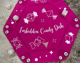 May include: A pink felt dice tray with white stitching and silver metal grommets. The tray features a white design of candy and the text "Forbidden Candy Dish".