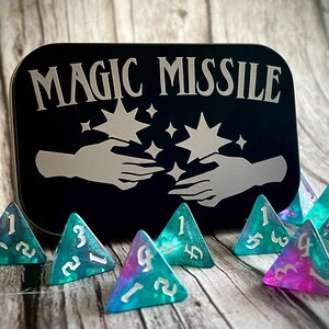 K&ouml;nnte beinhalten: Eine schwarze Metalldose mit der Aufschrift "MAGIC MISSILE" in silbernen Buchstaben. Die Dose ist mit zwei H&auml;nden verziert, die mit Sternen dar&uuml;ber nach oben reichen. Es gibt acht dreieckige, teal- und violettfarbene, schimmernde W&uuml;rfel mit wei&szlig;en Zahlen.