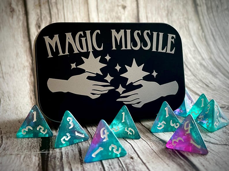 K&ouml;nnte beinhalten: Eine schwarze Metalldose mit der Aufschrift "MAGIC MISSILE" in silbernen Buchstaben. Die Dose ist mit zwei H&auml;nden verziert, die mit Sternen dar&uuml;ber nach oben reichen. Es gibt acht dreieckige, teal- und violettfarbene, schimmernde W&uuml;rfel mit wei&szlig;en Zahlen.