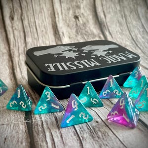 K&ouml;nnte beinhalten: Eine schwarze Metalldose mit der Aufschrift "Magic Missile" auf dem Deckel. Die Dose ist ge&ouml;ffnet und enth&auml;lt einen Satz von acht schillernden t&uuml;rkisfarbenen und violetten W&uuml;rfeln mit wei&szlig;en Zahlen.
