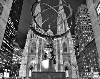 May include: Black and white photograph of the Atlas statue in front of St. Patrick's Cathedral in New York City. The statue depicts Atlas holding a large sphere, with the cathedral's spires rising in the background. The image is taken at night.