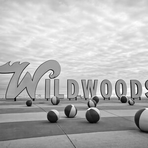 May include: Black and white photo of a sign that reads "WILDWOODS" with beach balls scattered on the ground in front of it.
