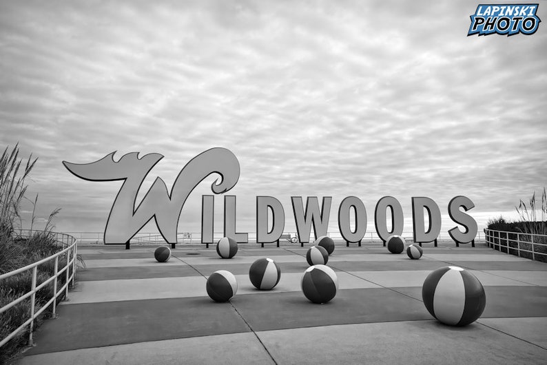 May include: Black and white photo of a sign that reads "WILDWOODS" with beach balls scattered on the ground in front of it.