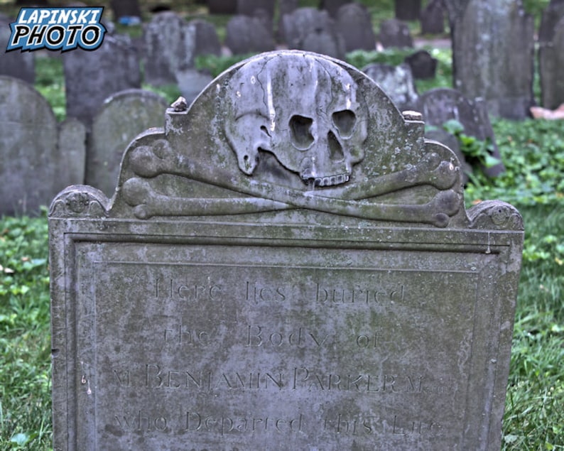 Puede incluir: Una l&aacute;pida gris y desgastada con una calavera y huesos cruzados tallados en la parte superior. La inscripci&oacute;n dice "Aqu&iacute; yace enterrado el cuerpo de BENJAMIN PARKER que parti&oacute; de esta vida".