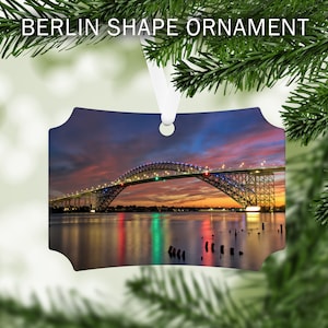 May include: A metal ornament featuring a photograph of a bridge lit up at night with a colorful sunset in the background. The bridge is reflected in the water below.
