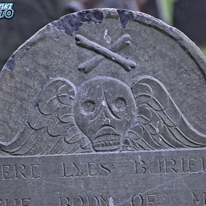 Puede incluir: Una l&aacute;pida de piedra gris desgastada con una calavera y huesos cruzados tallados. La inscripci&oacute;n dice "Here Lyes Buried The Body Of M..."