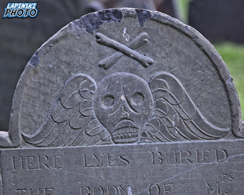 Puede incluir: Una l&aacute;pida de piedra gris desgastada con una calavera y huesos cruzados tallados. La inscripci&oacute;n dice "Here Lyes Buried The Body Of M..."