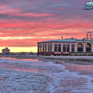 May include: A pink building with arched windows stands on the beach at sunset. The sky is a vibrant pink and orange. A Ferris wheel can be seen in the background.