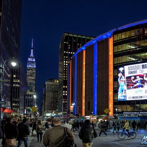 May include: A night view of Madison Square Garden in New York City with the Empire State Building in the background. The building is lit up in blue and orange lights. A large digital billboard advertises a basketball game with the text "Marquee Matchup Tonight 7:30".