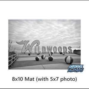 May include: Black and white photo of the iconic "Wildwoods" sign on the boardwalk with beach balls scattered in front of it. The sign is made of large, white letters with a black outline. The photo is taken from a low angle, looking up at the sign.