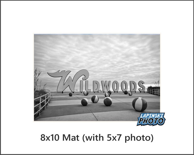 May include: Black and white photo of the iconic "Wildwoods" sign on the boardwalk with beach balls scattered in front of it. The sign is made of large, white letters with a black outline. The photo is taken from a low angle, looking up at the sign.