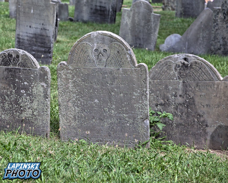 Puede incluir: Una fila de l&aacute;pidas grises y desgastadas en un cementerio. Las l&aacute;pidas est&aacute;n decoradas con calaveras y alas talladas. Las l&aacute;pidas est&aacute;n rodeadas de hierba verde.