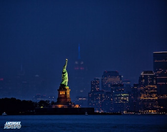 May include: A nighttime view of the Statue of Liberty illuminated against a dark blue sky with the New York City skyline in the background.