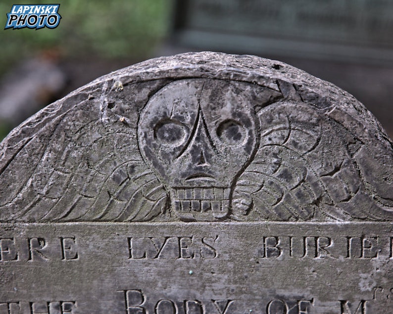 Puede incluir: Una l&aacute;pida de piedra gris desgastada con un dise&ntilde;o tallado de calavera y huesos cruzados. La inscripci&oacute;n dice "Here Lyes Buried The Body Of..."