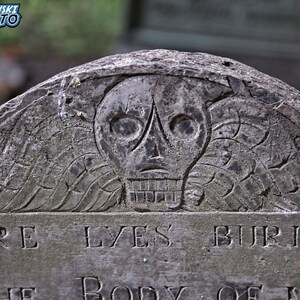 May include: A weathered gray stone tombstone with a carved skull and crossbones design. The inscription reads "Here Lyes Buried The Body Of..."