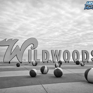May include: Black and white photo of a large sign that reads "Wildwoods" with beach balls scattered on the ground in front of it.