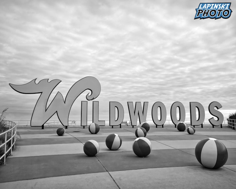 May include: Black and white photo of a large sign that reads "Wildwoods" with beach balls scattered on the ground in front of it.