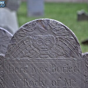 Puede incluir: Una l&aacute;pida de piedra gris desgastada con una inscripci&oacute;n que dice "Here lyes Buried ye Body of M's". La l&aacute;pida est&aacute; decorada con una calavera tallada y alas.