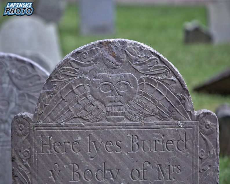 Puede incluir: Una l&aacute;pida de piedra gris desgastada con una inscripci&oacute;n que dice "Here lyes Buried ye Body of M's". La l&aacute;pida est&aacute; decorada con una calavera tallada y alas.