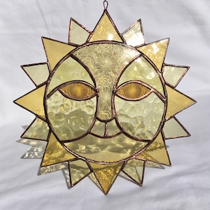 May include: A stained glass sun with a face, featuring yellow and brown glass. The sun has a textured surface and is framed in copper.